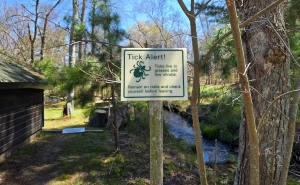 Near a wooded area of a park, a posted sign reads, Tick Alert! Ticks live in grasses and low shrubs. Remain on trails and check yourself before leaving.
